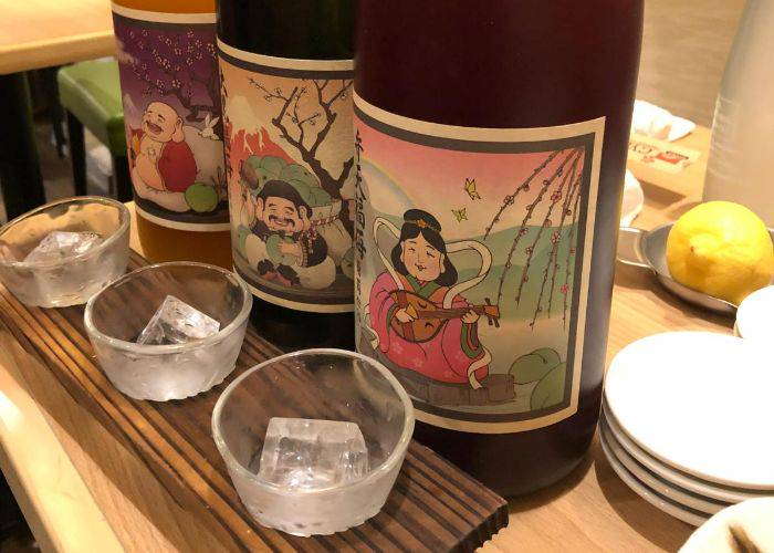 A sake tasting board with three cups, each with an ice cube. Behind them, three different sake bottles with eye-catching labels.